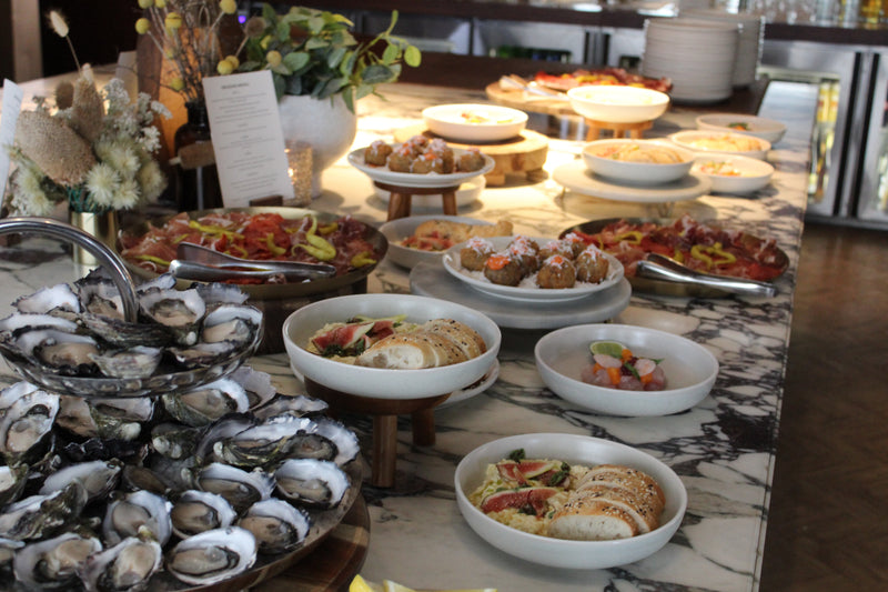 A marble buffet table featuring fresh oysters, charcuterie, bread with figs, and various appetizers in ceramic bowls.