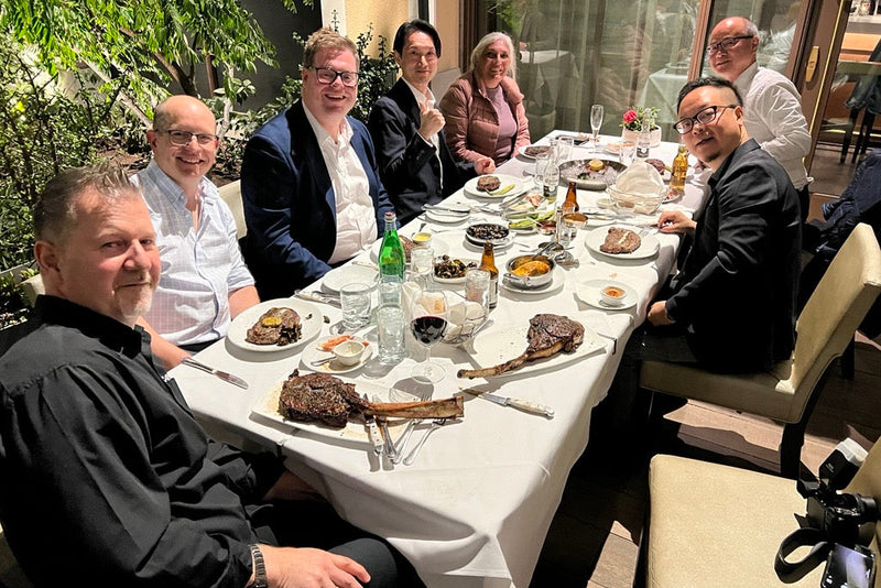 Group of colleagues enjoying a dinner with large tomahawk steaks at a white-clothed table on an outdoor patio.