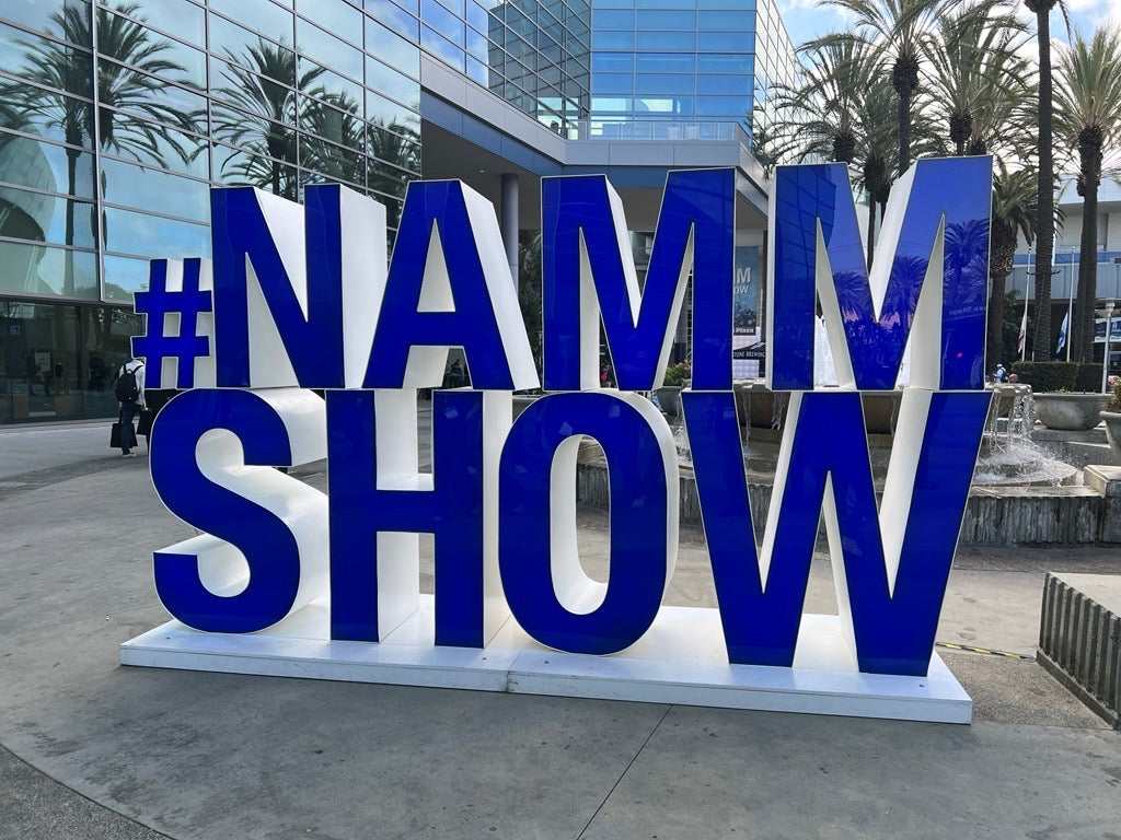 Large blue and white 3D #NAMM SHOW sign standing on a plaza in front of a modern glass convention center building.