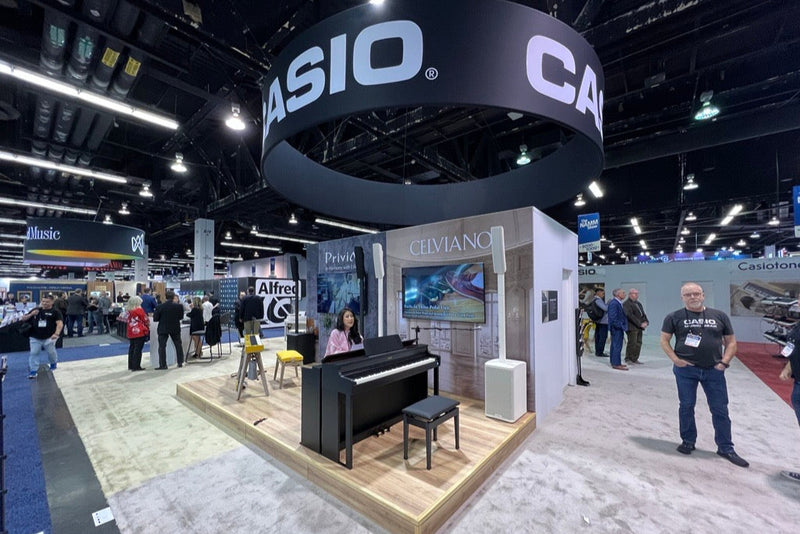 Casio trade show booth featuring a black digital piano, a woman playing, and a large circular hanging sign in a convention hall.