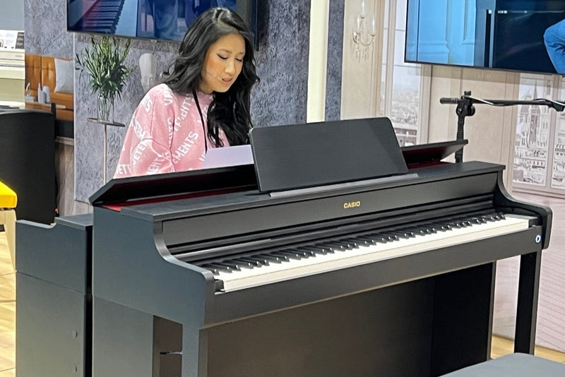 Woman in a pink sweatshirt playing a black Casio digital piano in a studio setting.