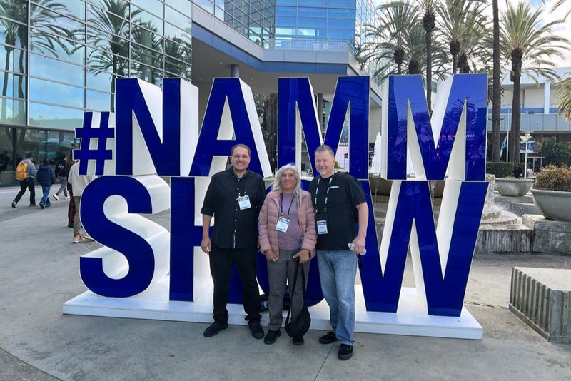 Three people posing in front of a large, blue and white #NAMM SHOW sign outside a convention center.