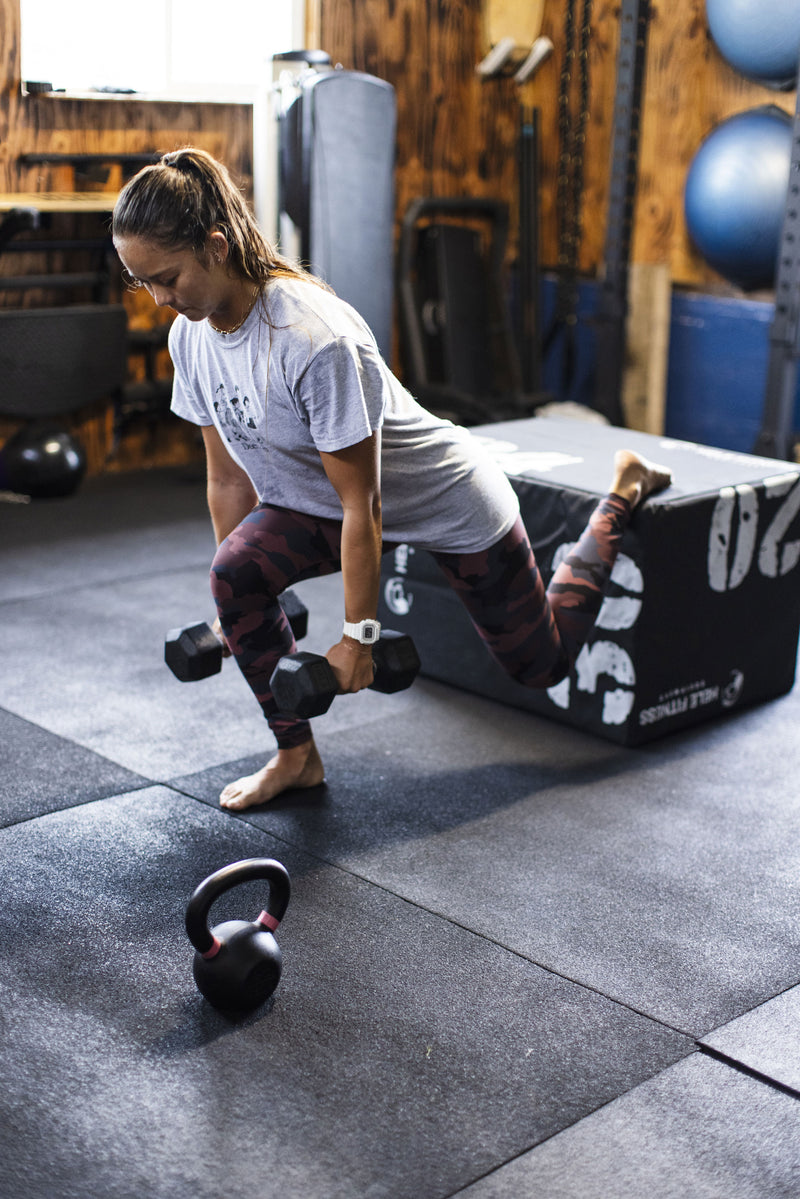Woman in a grey t-shirt and camo leggings performing a split squat with dumbbells in a gym with a kettlebell nearby.