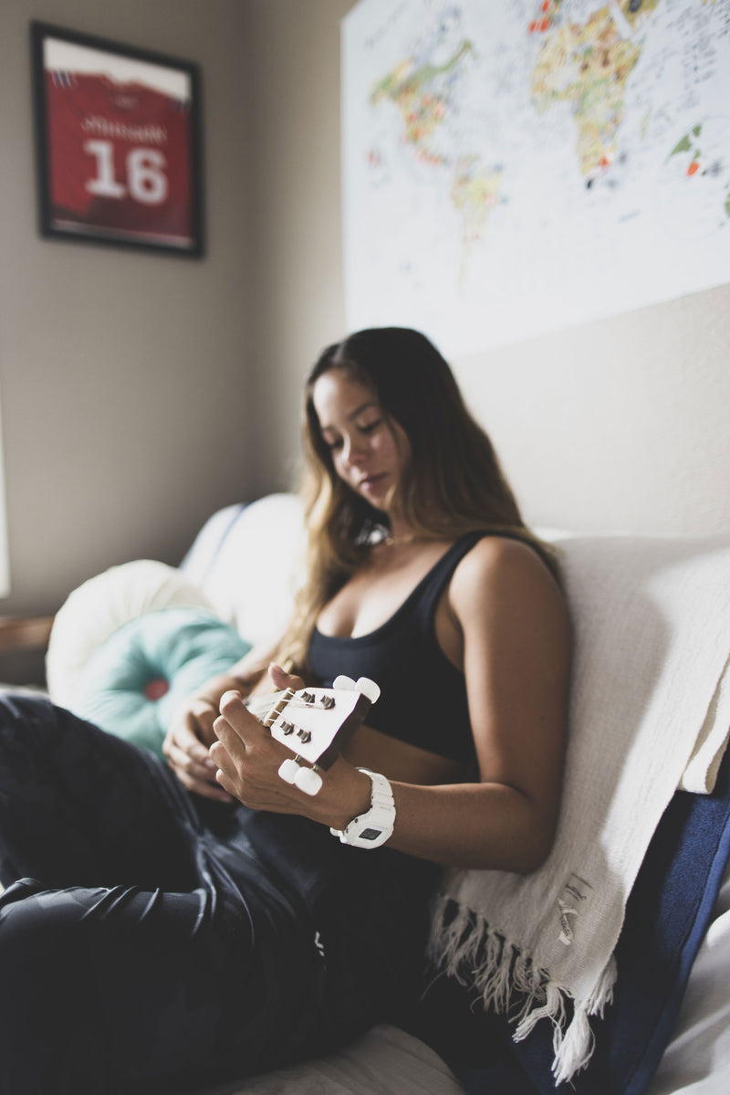 Young woman with long hair playing a light-colored ukulele while sitting on a couch with decorative pillows.