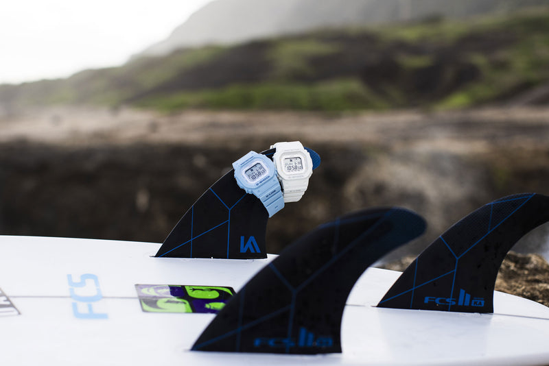 Light blue and white G-Shock G-LIDE digital watches resting on a surfboard fin at a beach.