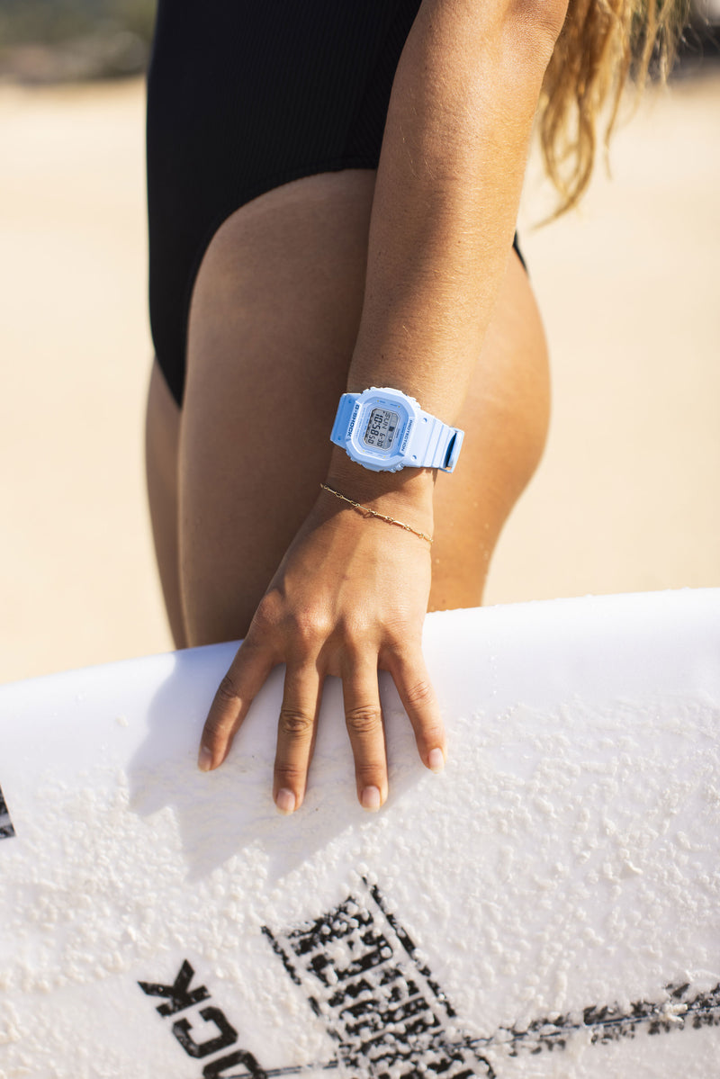 Person wearing a light blue G-Shock digital watch and gold bracelet while holding a white surfboard on a sandy beach.