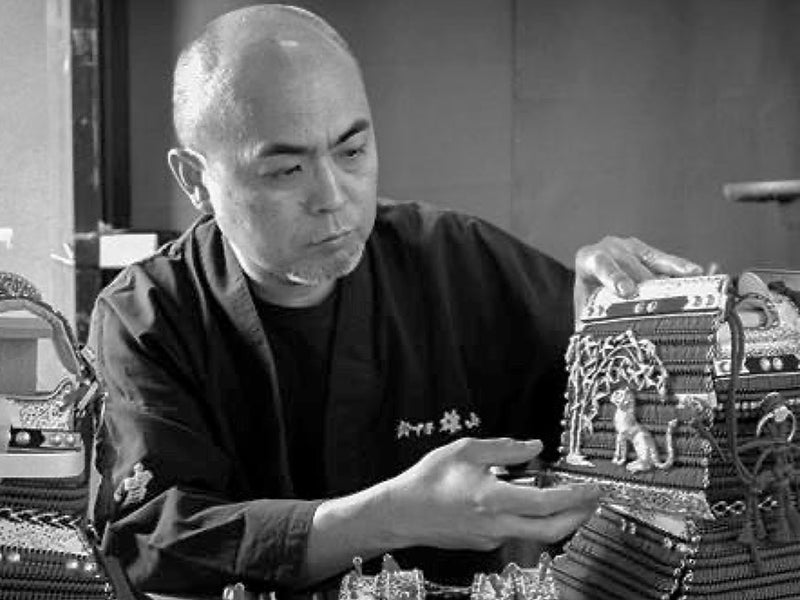 A Japanese craftsman in traditional attire meticulously assembling a detailed, ornate samurai armor piece.
