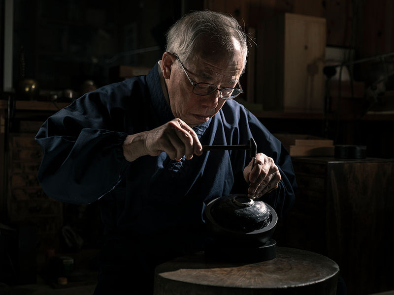 An elderly Japanese craftsman in a blue kimono uses a small hammer to meticulously detail a dark, circular metal object.
