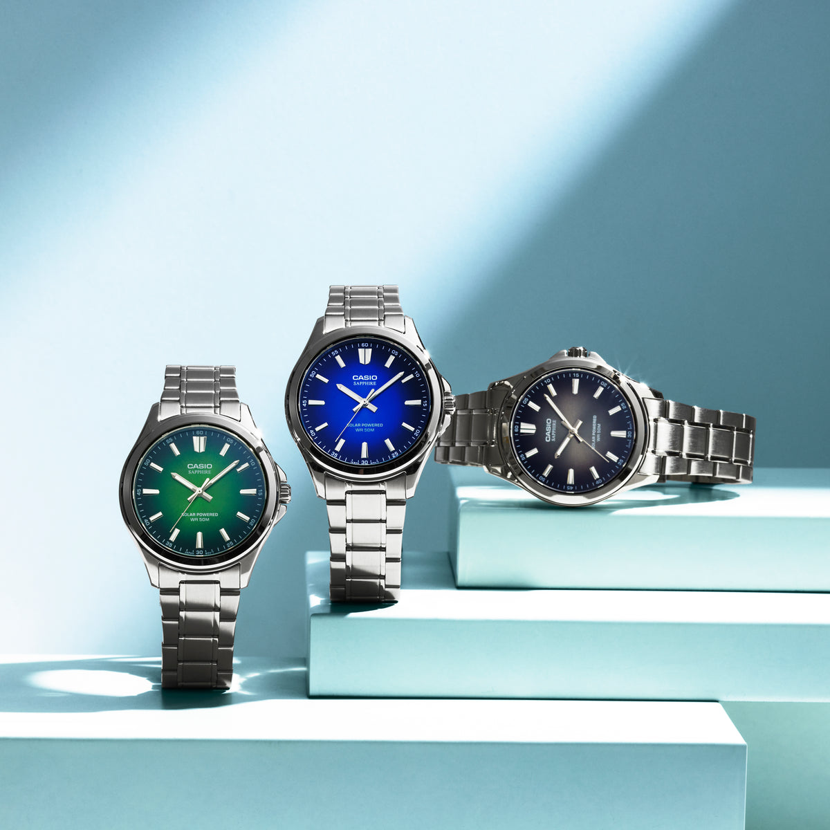 Three Casio solar-powered watches with stainless steel bands and gradient green, blue, and black dials on light blue pedestals.