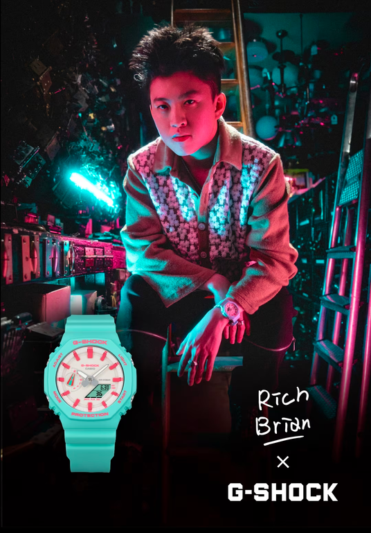 Rich Brian wearing a floral cardigan in a neon-lit studio next to a teal and pink G-Shock watch.
