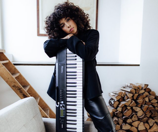 Woman with curly hair in a black blazer leaning on a vertical Casio keyboard in a rustic, modern living space.