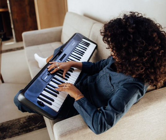 A person with curly hair sits on a beige couch playing a black Casio electronic keyboard.