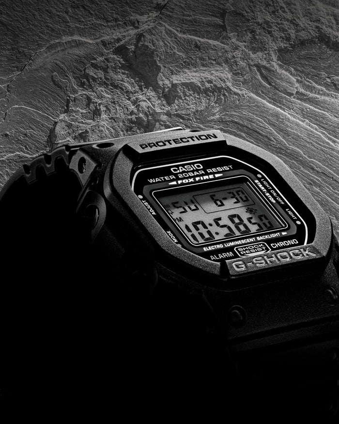 Black Casio G-Shock digital watch with a square face and resin strap set against a textured, dark rocky background.