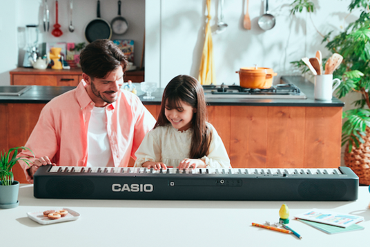A father and daughter playing a black Casio CDP-S160 digital piano together at a kitchen table.