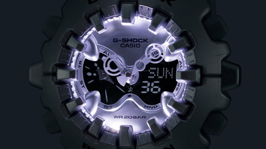 Close-up of a black G-Shock watch face with illuminated silver hands, digital display, and rugged gear-shaped bezel.