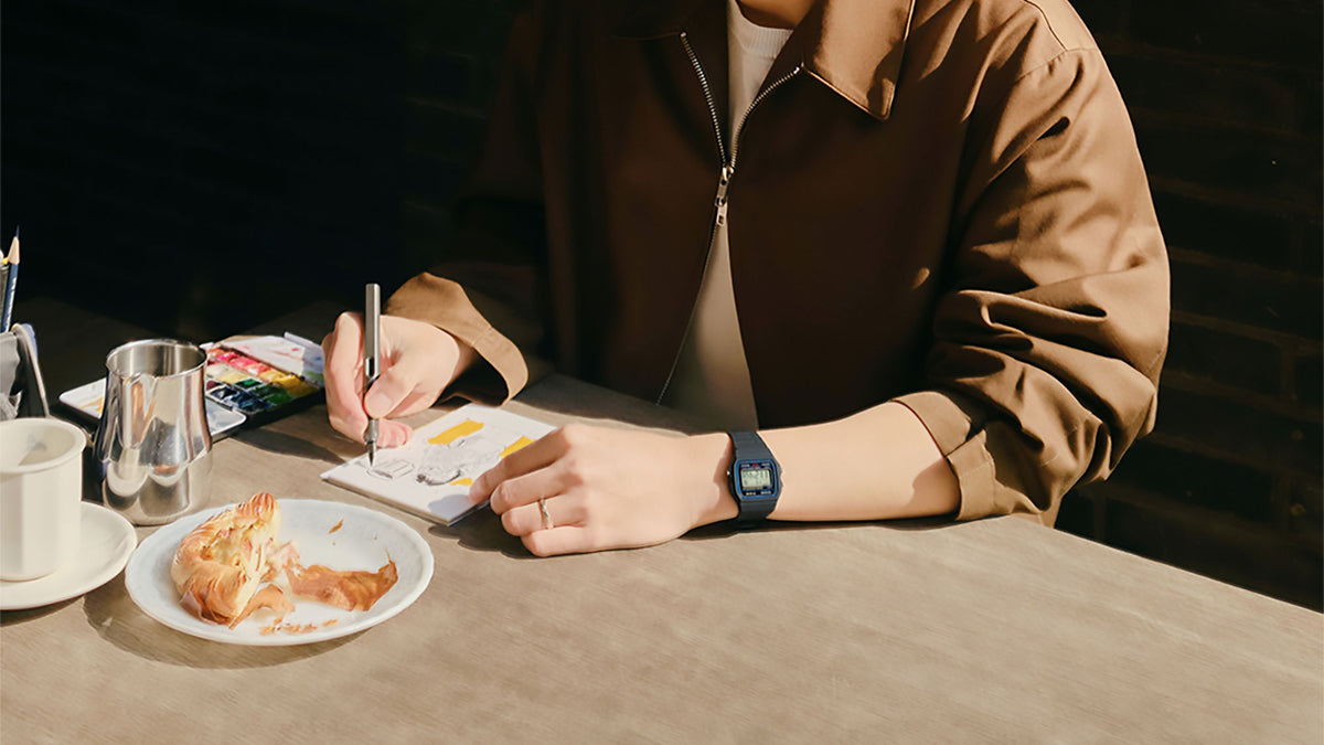 Person in a brown jacket sketching at a table while wearing a classic black Casio F-91W digital watch.