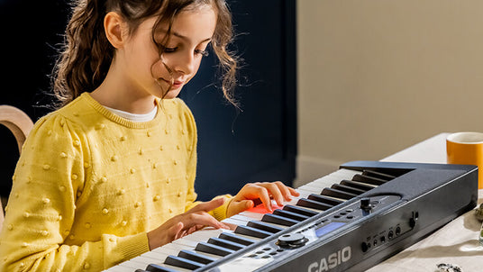 Young girl in a yellow sweater playing a black Casio keyboard with illuminated keys at a table.