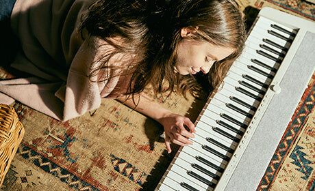 Young woman with long hair lying on a patterned rug playing a slim, modern white digital keyboard.