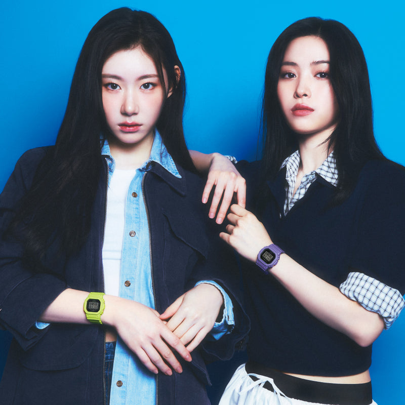ITZY members posing against a blue background wearing Casio G-Shock watches in lime green and purple.