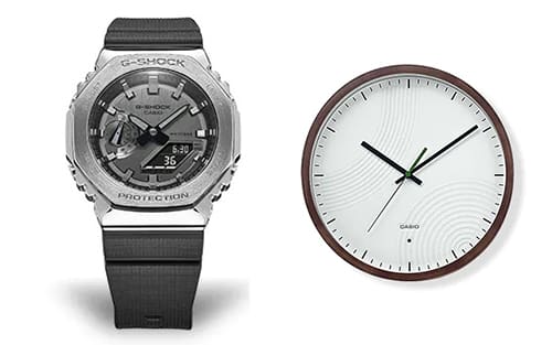 A silver Casio G-Shock wristwatch with a black strap next to a round white wall clock with a dark wooden frame.