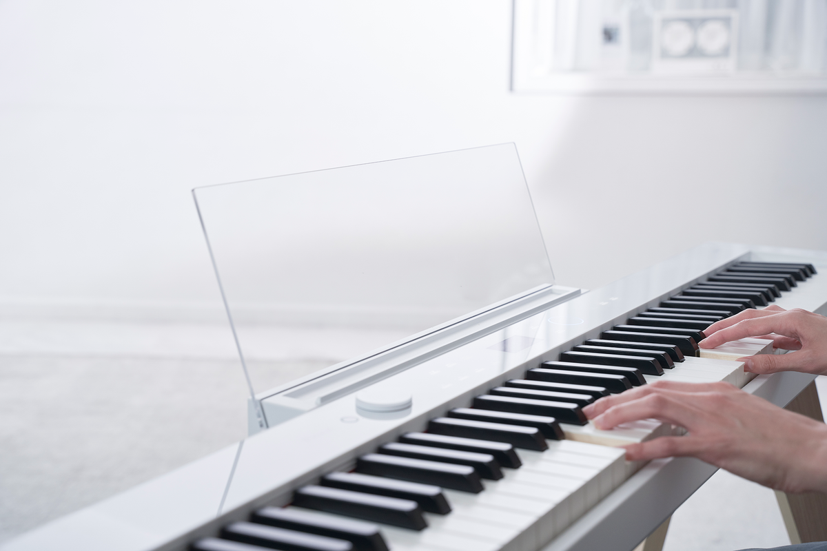 Hands playing a sleek white Casio Privia PX-S7000 digital piano with a transparent music rest in a bright room.