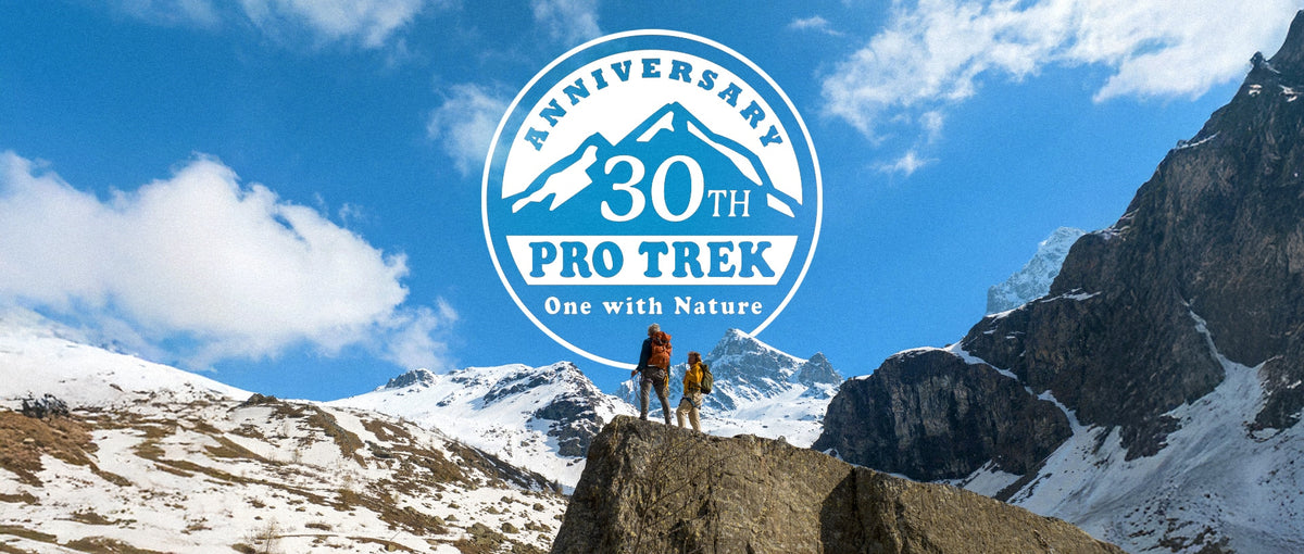 Two hikers standing on a rocky peak before snow-capped mountains with a blue Pro Trek 30th anniversary logo overlay.